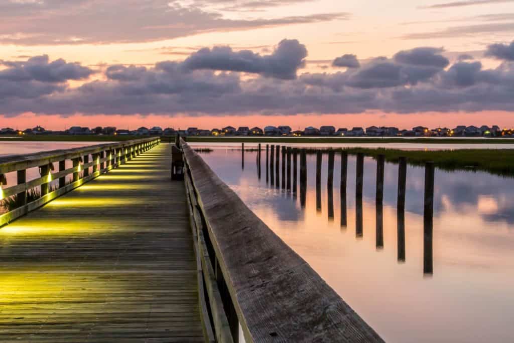 View down the Marshwalk on Murrells Inlet toward the horizon.. The Marshwalk is a wooden bridge and is lit by yellow spotlights on the left side of the walk. On the right are wooden posts and their reflection in the water. One the horison are houses, then a pink sky, then puffy white clouds.