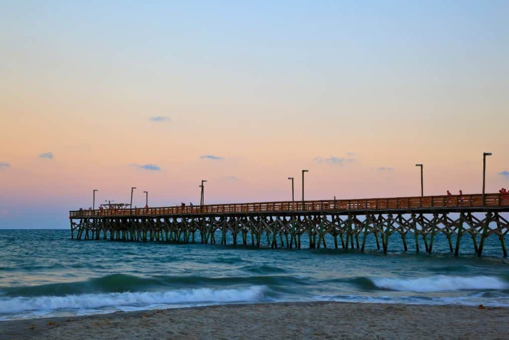 Surfside Pier in South Carolina. Pier extends into the ocean from right to left. Early morning, sky is blue, pink and yellow with a few small puffy clouds. Ocean waves are small and break on the beach. A strip of sand is visible at the bottom of the photo.