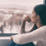 Teenage girl in pink fully sweater with long dark hair looking out the window in profile. Her knees are pulled up and one arm hugs her knees while the other rests on her knee with her hand at her mouth. She looks sad and lonely.
