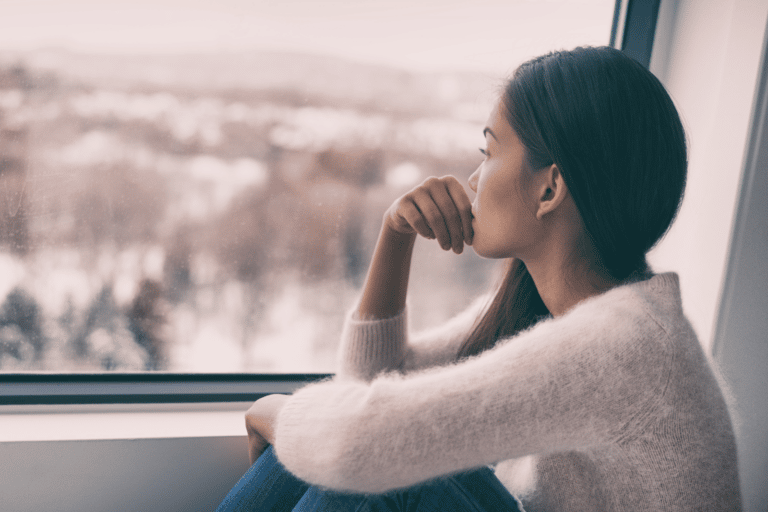 Teenage girl in pink fully sweater with long dark hair looking out the window in profile. Her knees are pulled up and one arm hugs her knees while the other rests on her knee with her hand at her mouth. She looks sad and lonely.