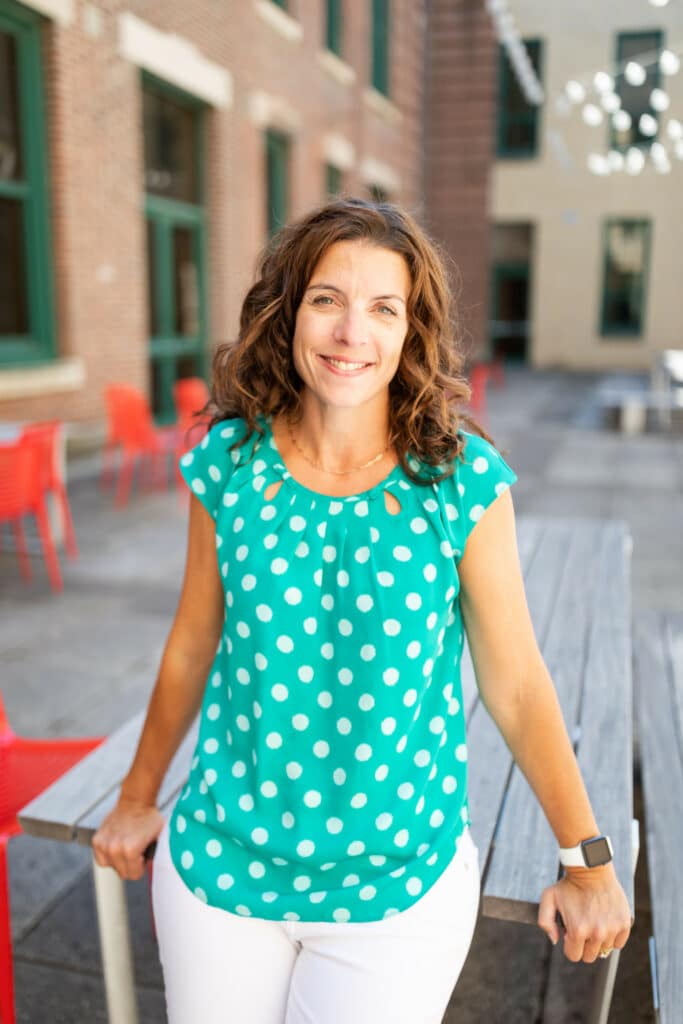Picture of woman leaning against a table. Dark hair, smiling, wearing a teal short sleeve blouse with white polka dots.