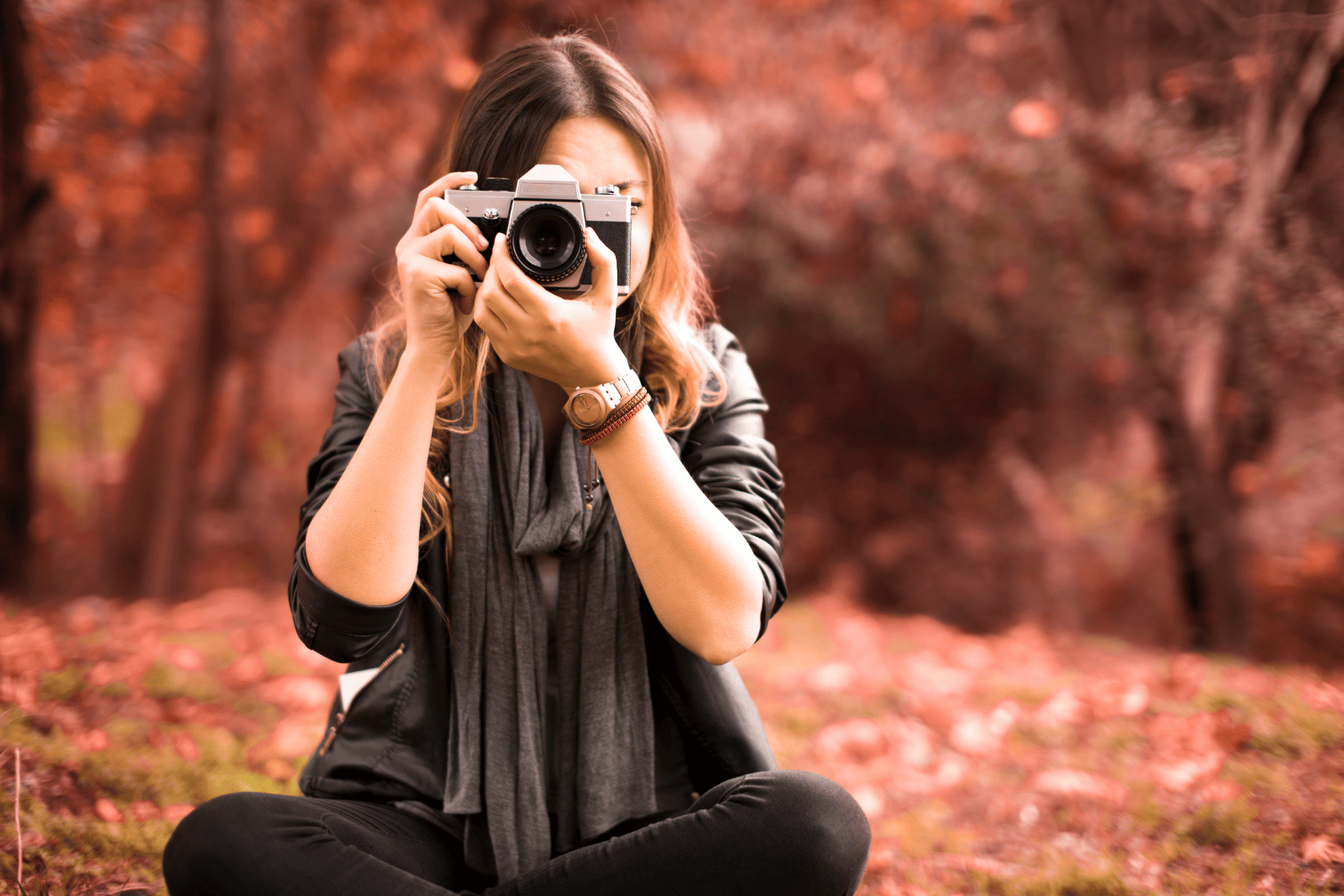 Woman facing the camera with a point and shoot camera in front of her face. She's wearing black pants, black jacket and a gray scarf, and is seated on the ground. Blurred fall leaves are behind her.