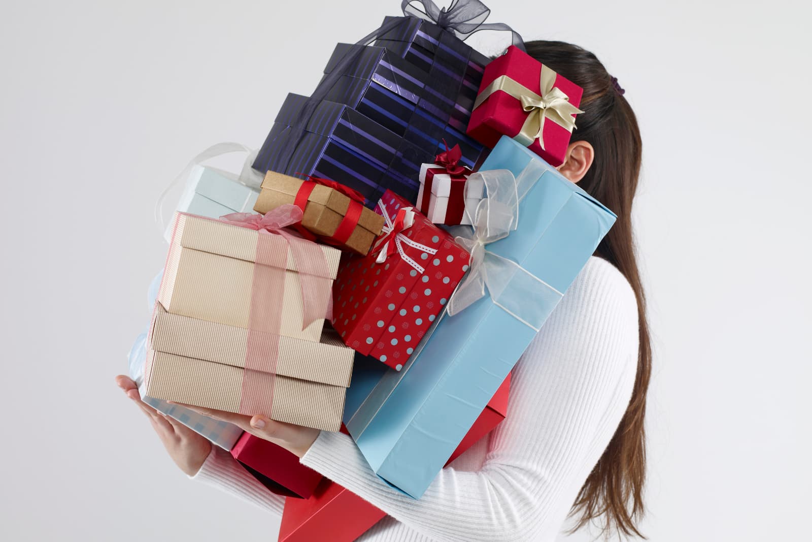 Teenage girl with long dark hair and long sleeve white sweater is balancing a large pile of gifts in her arms. Gifts are varied colors of boxes and bows.