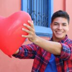Dark haired older teenage boy wearing a red and blue plaid shirt with sleeves rolled up and blue shirt underneath. Holding a heart shaped balloon with extended arms toward camera. Standing in front of a reddish building with blue windows with iron bars.