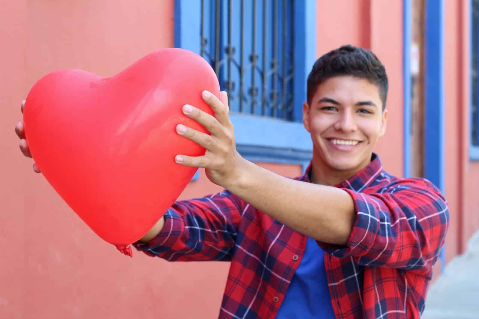 Dark haired older teenage boy wearing a red and blue plaid shirt with sleeves rolled up and blue shirt underneath. Holding a heart shaped balloon with extended arms toward camera. Standing in front of a reddish building with blue windows with iron bars.