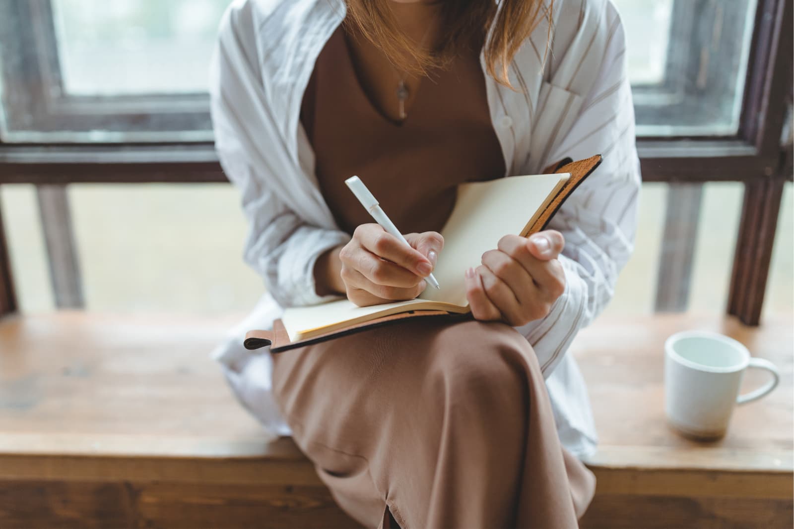 woman in brown dress with white long sleeve shirt over top, sitting on a window sill writing in an open journal with a white pen. A white coffee mug sits on the sill on the right side of the image