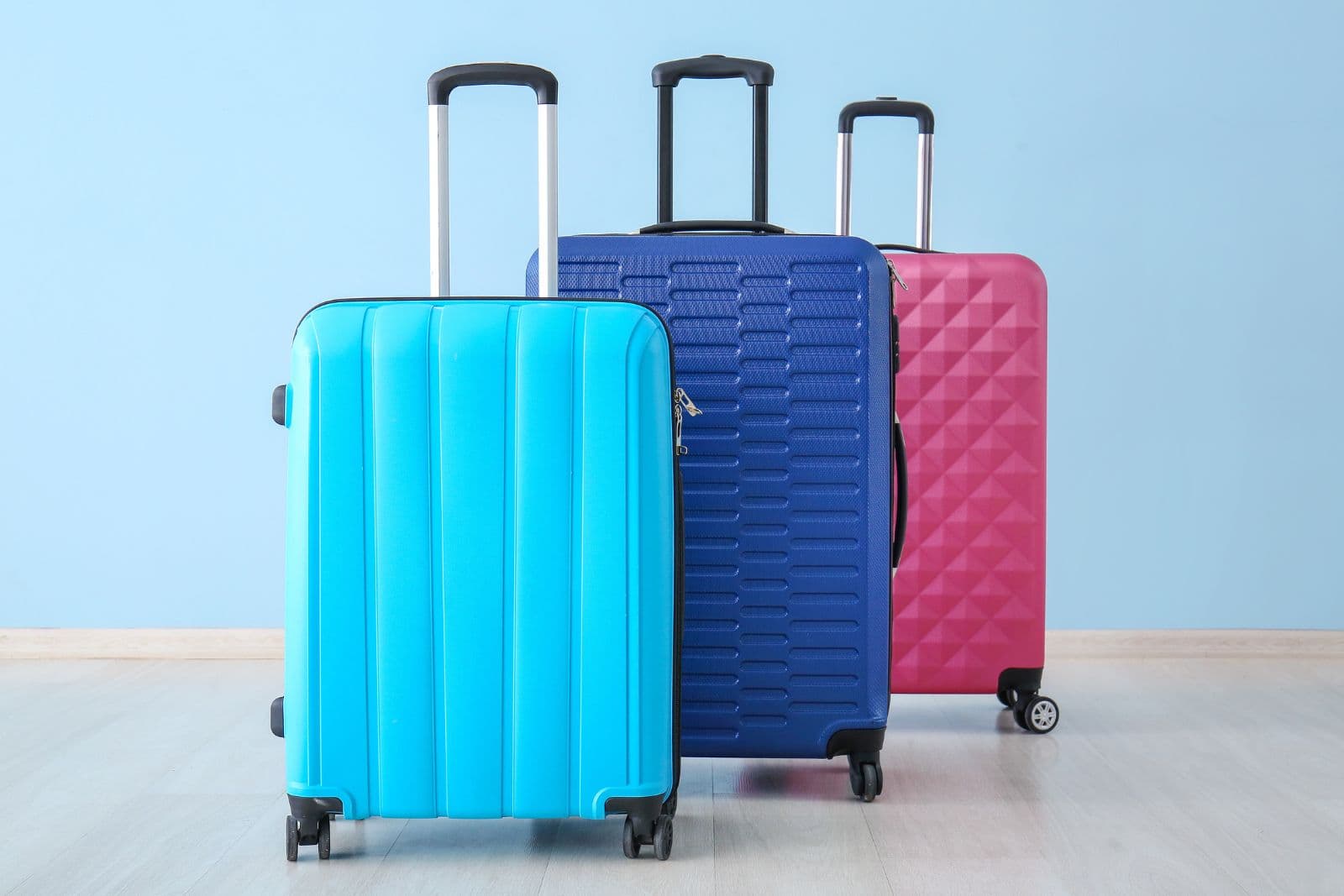 Hardshell luggage in trendy teen colors and patterns against a light blue wall. Three suitcases are offset from each other, one is teal with wide vertical lines, one is navy with a horizontal line pattern and one is fuscia with a diamond pattern. All have wheels and extended handles