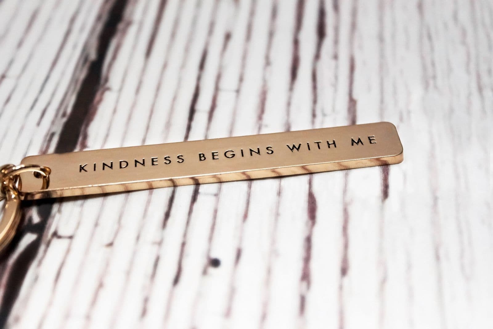 Rose gold keychain that says "kindness begins with me" on a white wood background.