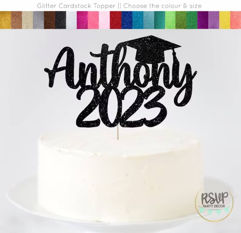 White cake on a white plate with a black glitter cake topper that says "Anthony 2023" with a graduation cap.