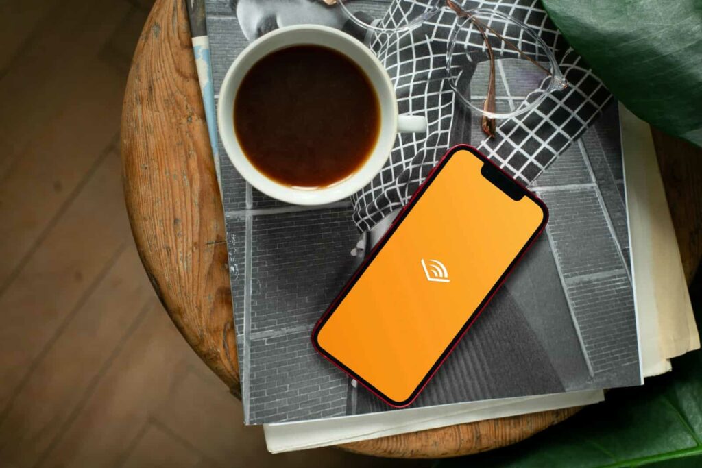 Overhead image of a stack of papers, a pair of glasses, a cup of tea and a smartphone on top of a wooden stool. The yellow Audible app is visible on the phone. Green leaves are visible on the right side of the stool