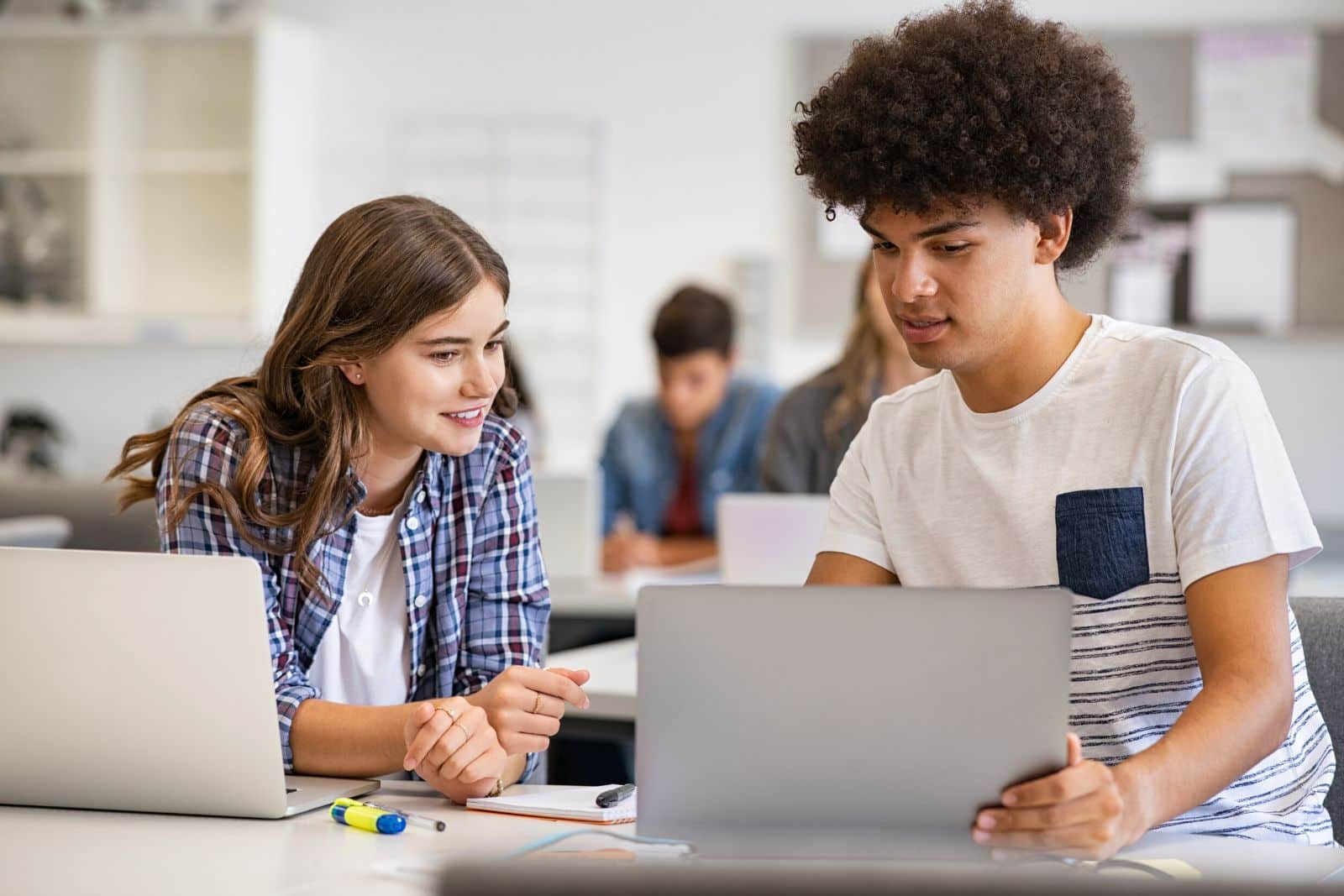Teenagers in a classroom sharing a table. A girl with long brown hair in a plaid shirt is leaning across her open laptop to a boy with black hair in a white shirt with blue stripes. The boy is pointing to something on his open laptop.