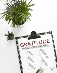 Flat lay on white background of a plant and black clipboard with white lines. On the clipboard is a paper that reads "Gratitude Photo Scavenger Hunt"