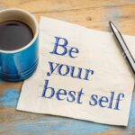 Flat lay of a blue mug of coffee, a napkin and a pen on a rustic wooden background with streaks of light blue paint. "Be your best self" is sketched in blue pen on the napkin.