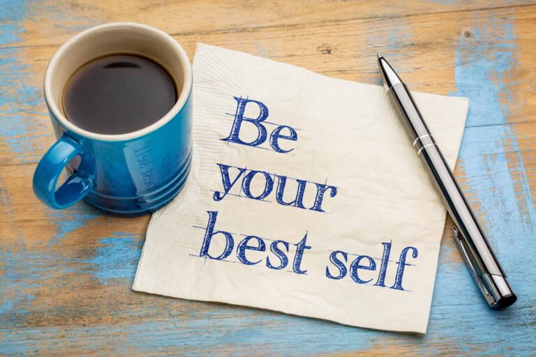Flat lay of a blue mug of coffee, a napkin and a pen on a rustic wooden background with streaks of light blue paint. "Be your best self" is sketched in blue pen on the napkin.