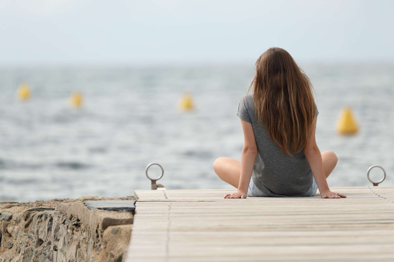 Teen girl with long brown hair sits on a pier looking out at the ocean. She's facing away from the camera, wearing a gray tee shirt and denim shorts.