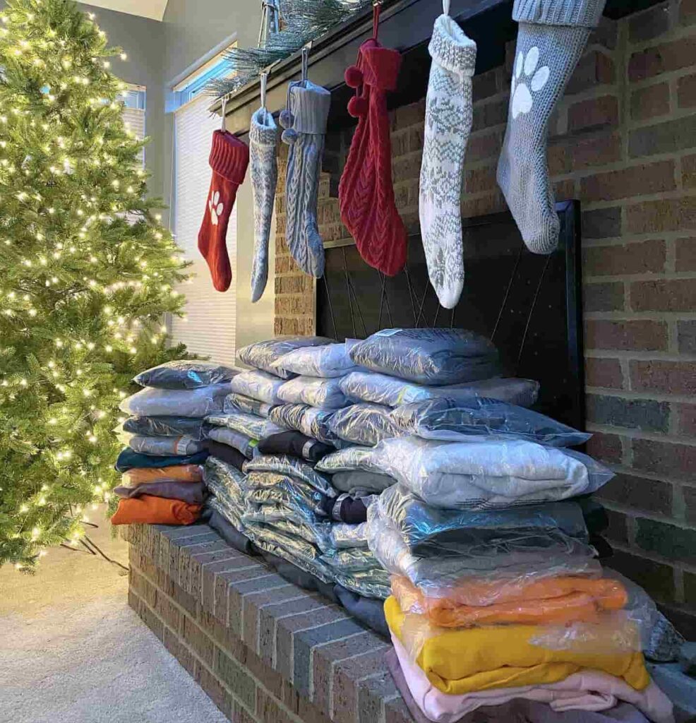 Photo of a fireplace decorated with stockings and garland for Christmas. Five stacks of hoodies cover the hearth. A Christmas tree with white lights in on the back left of the fireplace and image.